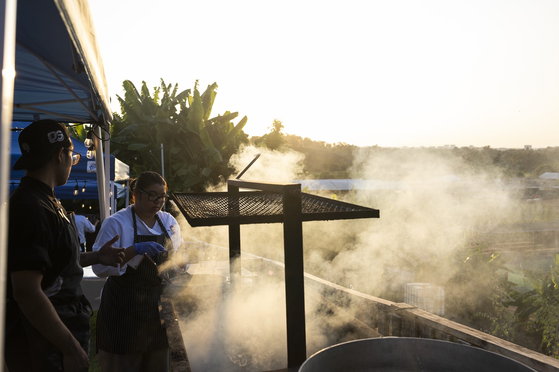 A chef grilling at sunset amid rising steam at Harvest Moon